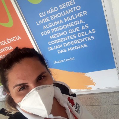 Foto de Talita Mendes. Ela está de máscara olhando para a frente com um semblante de alegria. Atrás dela há um painel com a seguinte frase: Eu não serei livre enquanto alguma mulher for prisioneira, mesmo que as correntes dela sejam diferentes das minhas, de Audre Lorde